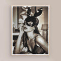 Framed artwork of a woman wearing a black mask with bunny ears on a beige wall with natural/oak frame